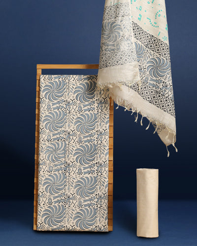 Off white and slate blue Katan silk unstitched suit set hand block printed with stylised swirls and fish motifs in an asymmetric repeat, with teal and charcoal grey accents and fringed edges.