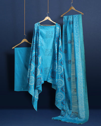 Sky blue Katan silk unstitched suit set block printed with tribal figures and circular mandalas in bright and light blue on the top piece, a dupatta bordered with linear geometric bands, and a plain bottom piece.