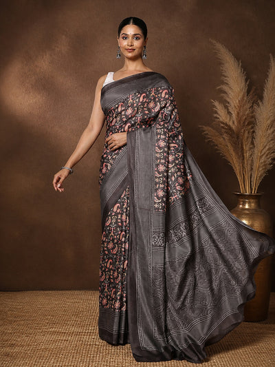 Charcoal grey printed saree with a floral and paisley print in pink, mustard and green, framed by a wide decorative border.